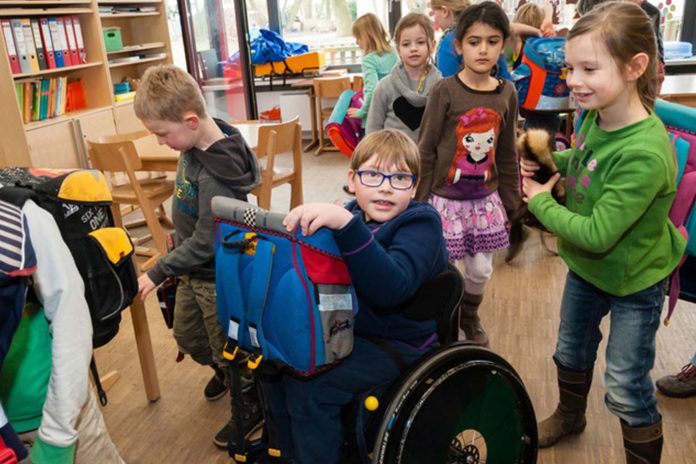 Immagine che ritrae bambini di una scuola primaria in classe, con giocattoli e zaini. Uno di loro, centrale nell'immagine, è su una sedia a rotelle con in braccio il suo zainetto